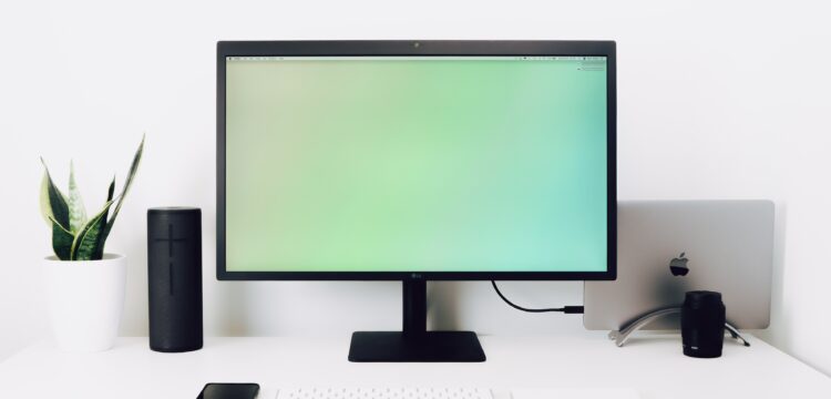 decorative image: a work desk with a big monitor, speakers, a keyboard and a mobile phone. In the back is a laptop stashed away and on the left you can see a plant.