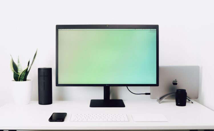 decorative image: a work desk with a big monitor, speakers, a keyboard and a mobile phone. In the back is a laptop stashed away and on the left you can see a plant.